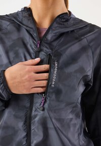 Giacca leggera nera con cappuccio, dotata di tasca con cerniera con dettagli viola e un logo sul davanti. Tessuto liscio e riflettente.