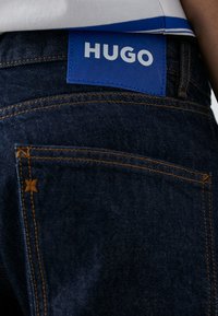 Jeans in denim blu scuro con cuciture arancioni e una patch in pelle blu con il marchio "HUGO" sulla cintura posteriore.