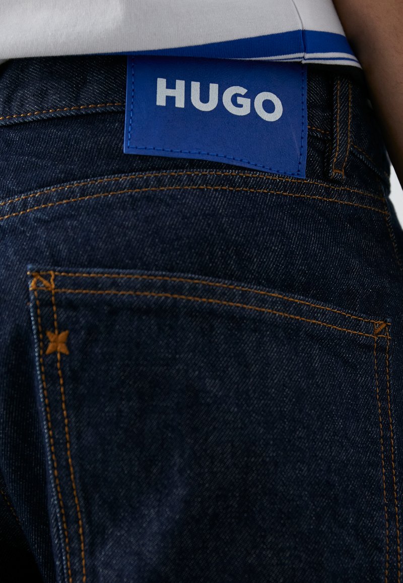 Granatowe dżinsy z pomarańczowymi przeszyciami i niebieską skórzaną metką z napisem "HUGO" na tylnej części pasa.
