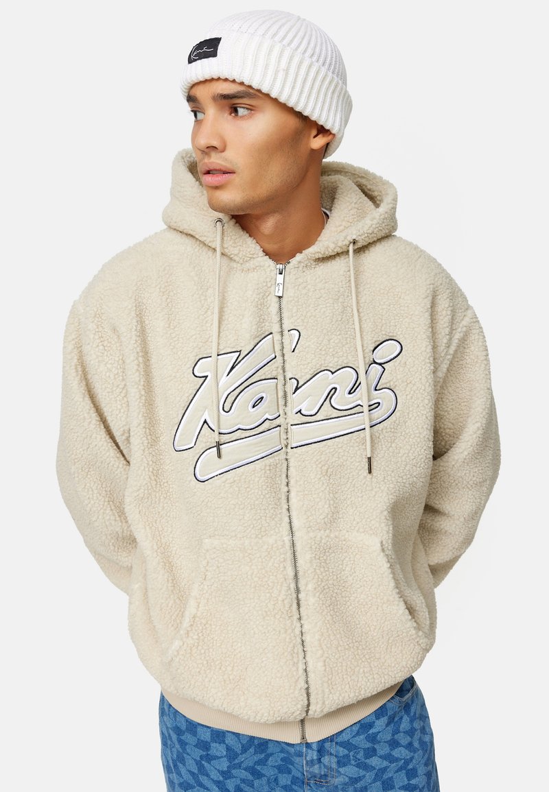 Junger Mann trägt einen beigen, flauschigen Kapuzenpullover mit Reißverschluss und dem Schriftzug „Karni“ sowie eine weiße Strickmütze, schaut nach links und hat die Hände in den Taschen.