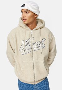 Hoodie in pile beige con zip frontale, grandi tasche e logo ricamato. Accompagnato da un berretto a costine bianco. Vestibilità standard.