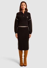 Czarny zestaw swetra i spódnicy z prążkowanej dzianiny z kremowymi bocznymi paskami, z kołnierzowym designem i łatką z logo. Modelka nosi brązowe kozaki za kolano.