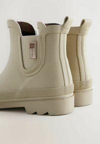 Bottines en caoutchouc beiges, avec une semelle texturée, des panneaux latéraux élastiques et une doublure à carreaux. Présente une étiquette de marque sur le côté latéral.