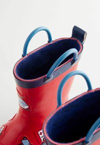 Bottes de pluie en caoutchouc rouge avec des poignées bleues, une doublure marine et des patchs décoratifs sur l'extérieur. Texture lisse avec une forme arrondie.