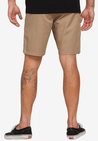 Beige Shorts mit flachen Taschen vorne und hinten, aus glattem Stoff. Getragen mit schwarzen Leinenschuhen. Sichtbares Oktopus-Tattoo am linken Bein.
