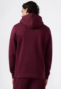 Felpa borgogna con tasca a marsupio, polsini e orlo a costine. Tessuto liscio con cappuccio regolabile, mostrata da dietro con capelli ricci visibili.