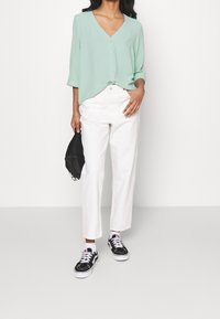 Blouse vert clair à col en V avec boutons et manches trois-quarts, associée à un pantalon blanc droit et des baskets noires. Sac noir à la main.
