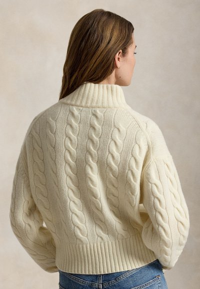 Kvinde med brunt hår iført en cremefarvet kabelstrikket rullekrave og blå jeans, set bagfra mod en simpel neutral baggrund.