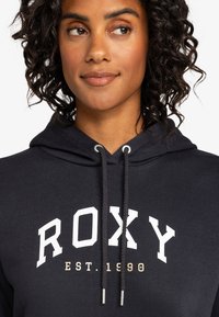 Dark navy hoodie with a white "ROXY" logo and "EST. 1990" in gold. Features a drawstring hood and soft fabric texture.