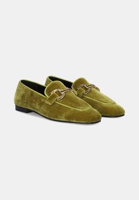 Par de mocasines de terciopelo verde oliva con detalle de cadena dorada en la parte superior, suelas negras y tacones bajos, mostrados sobre un fondo claro y liso.