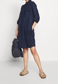 Navy-blaues Hemdblusenkleid aus leichtem Stoff, mit zwei Brusttaschen, hochrollbaren Ärmeln und Seitentaschen. Kombiniert mit beigen Sandalen und einem grauen Rucksack.