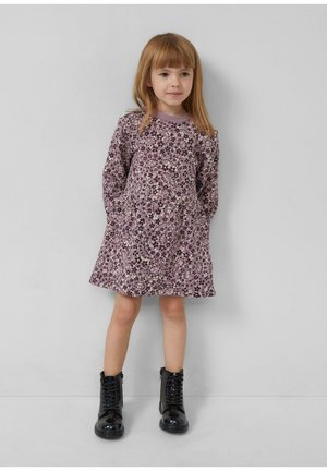 Junges Mädchen mit glattem, lichtbraunem Haar, das ein langärmeliges lila Blumenkleid und schwarze glänzende Schnürstiefel trägt, steht an einer grauen Wand.