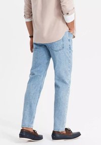 Jeans de mezclilla azul claro con un corte relajado, dos bolsillos traseros y una textura desgastada, combinados con zapatos casuales oscuros y una camisa beige.