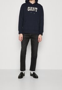 Persona con una sudadera azul marino que lleva el texto "GIANT", jeans ajustados negros, calcetines blancos y mocasines negros, de pie contra un fondo liso.