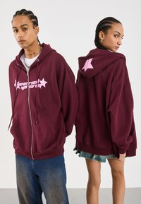 Hoodie zippé bordeaux en tissu doux, avec du texte rose et des graphismes d'étoiles sur le devant et le dos. Coupe ample avec poches avant.