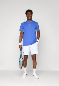 Male tennis player wearing blue and green shirt, white shorts, white wristbands, holding black and blue tennis racket, standing against plain background.