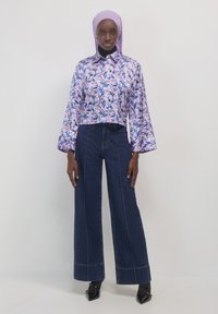 Frau in einer lavendelfarbenen Bluse mit Blumenmuster, hellem lila Hijab, hoch taillierten, weit geschnittenen dunklen Jeans und schwarzen spitzen Schuhen, stehend vor einer weißen Wand.