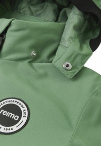 Giacca per bambini verde impermeabile, con un'esterno liscio, fodera interna trapuntata, chiusura a scatto e un patch circolare con logo.