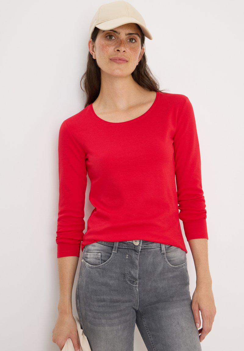 Röd långärmad skjorta i mjukt material med rundad halsringning, kombinerad med urtvättade grå jeans och håller en ljus färgad kuvertväska.