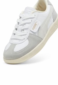 Puma Palermo sneakers har en ovandel av vit läder med ljusgrå mockadetaljer, platta skosnören och en texturerad krämfärgad gummisula.