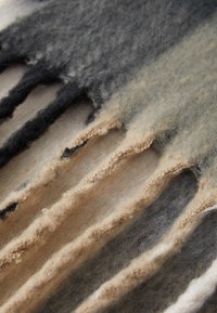 Tissu en laine texturé avec des tons beige, noir et gris. Présente des bords irréguliers et un design superposé et ondulé avec une texture douce et duveteuse.