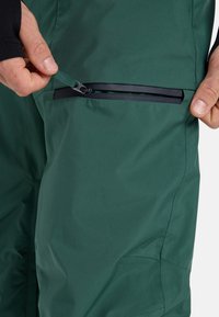I pantaloni impermeabili verdi presentano una tasca con zip e un dettaglio della zip nera. Il tessuto ha una struttura liscia e un design aderente.
