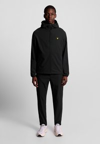 Veste légère noire avec capuche, logo jaune sur la poitrine, portée avec un pantalon noir assorti et des chaussures de sport claires. Texture lisse, design épuré.