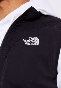 Nahaufnahme einer Person, die eine schwarze Jacke mit Reißverschluss trägt, auf der die gestickte Marke "The North Face" auf der Brust über einem weißen Hoodie zu sehen ist.