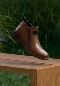 Braune Leder-Stiefelette mit einem seitlichen Reißverschluss und einem strukturierten schwarzen Akzent. Glatte Oberfläche und runde Zehenpartie; robuste Gummisohle für guten Halt.