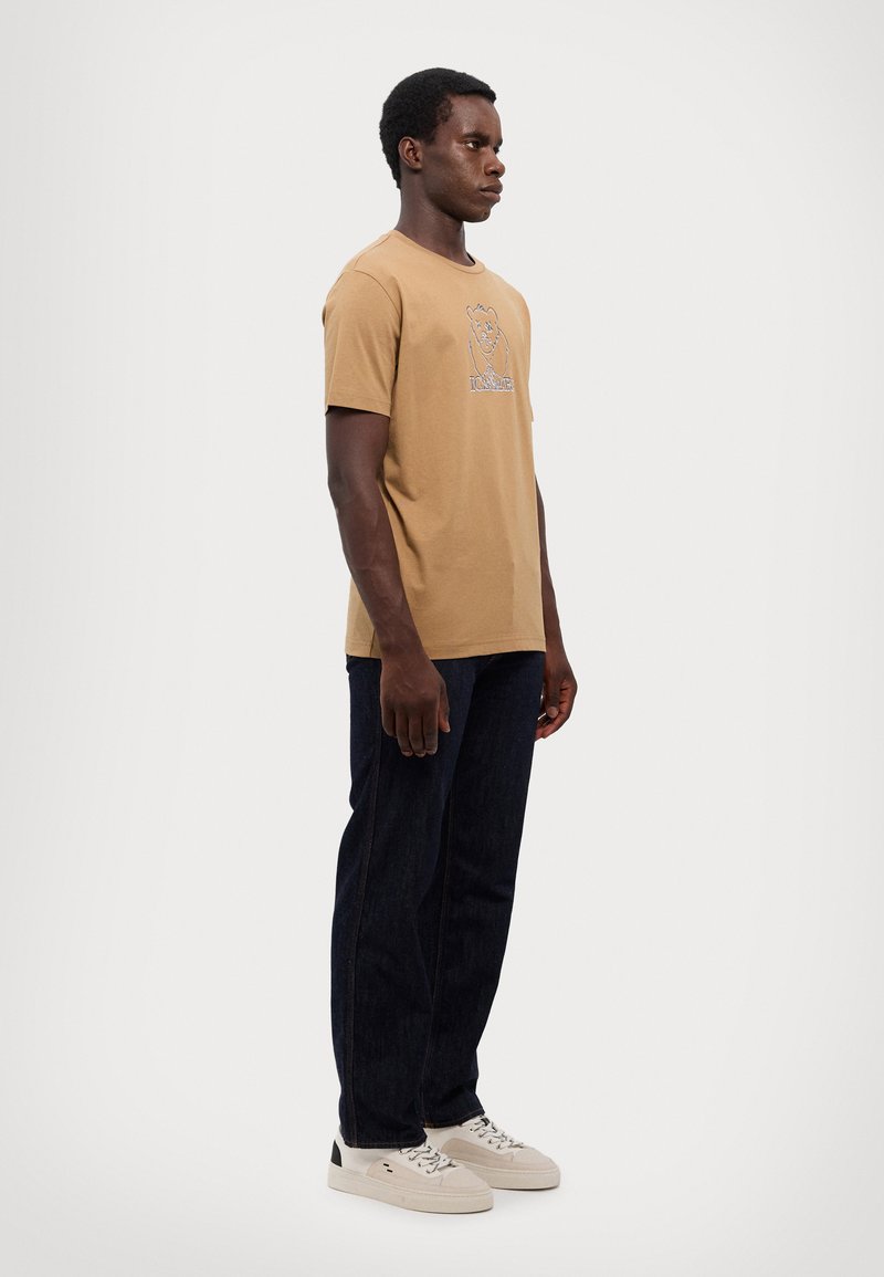 T-shirt marron à manches courtes avec un graphique d'ours ; associé à un jean bleu foncé et des baskets blanches. Le t-shirt est en tissu coton doux.