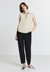 Top beige senza maniche con colletto a punta, abbinato a pantaloni neri a sigaretta e scarpe slingback argentate, su uno sfondo neutro.