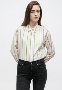 Hosszú ujjú ing világos rózsaszín és zöld függőleges csíkokkal, patentos eleje, klasszikus gallérral, sötét fekete farmernadrággal viselve.