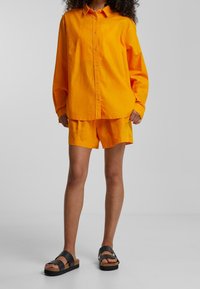 Orange bomullsskjorta med knappar och långa ärmar, matchad med orange shorts. Modellen bär svarta sandaler med korksålar.