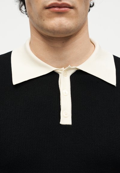 Κοντινό πλάνο ενός ατόμου που φοράει μαύρο πλεκτό πόλο με λευκό γιακά και τρία λευκά κουμπιά, κομμένο κάτω από τη μύτη.