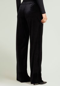 Zwarte, wijde broekspijpen gemaakt van zacht, fluweelachtig materiaal met een gladde textuur en een hoge tailleband. De stof valt elegant aan de achterkant.