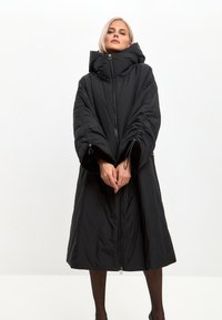 Sort oversized pufferjakke med høj krave, lynlås fortil og elastiske manchetter, der har en glat, polstret tekstur og et mellemlangt design.