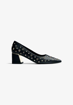 Zapato de piel negra con punta afilada, adornado con tachuelas metálicas doradas, y un tacón en bloque geométrico que presenta una franja dorada.