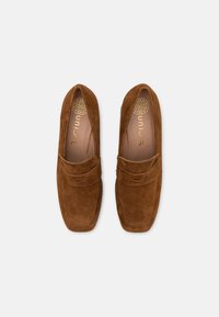 Hnědé semišové mokasíny s plochým designem, které mají dekorativní pásek přes svršek a hladkou koženou stélku. Zlaté logo uvnitř.