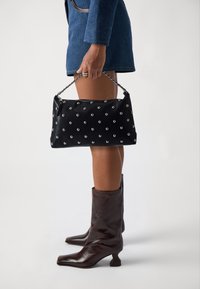 Sac à main noir avec des clous en argent, en forme rectangulaire, en matériau texturé, et avec une poignée en chaîne. Associé à des bottes marron à bout carré.