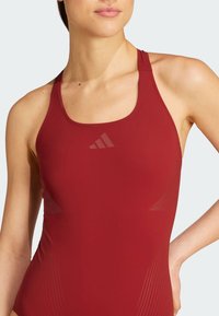 Red athletic swimsuit with a scoop neckline, thin straps, and subtle line patterns along the sides. Features a transparent logo accent.