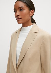 Beżowy żakiet z klapami, teksturowana tkanina i dopasowany krój, zestawiony z białą bluzką o wysokim szyi z prążkami. Prosty i elegancki design.