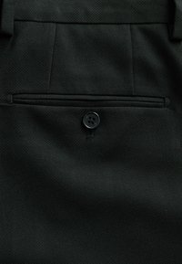 Tissu noir avec un seul bouton fermant une poche arrière horizontale sur un pantalon tailleur avec passants de ceinture.