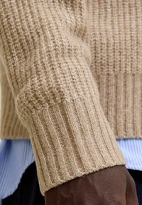 Beige knitted sweater featuring ribbed texture, soft material, and a wide cuff, layered over a blue and white striped shirt.