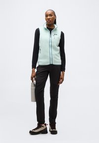 Γυναίκα που στέκεται φορώντας ορειβατικά μποτάκια, μαύρο παντελόνι, μαύρο μακρυμάνικο μπλουζάκι, ανοιχτό μπλε γιλέκο από φλις, κρατώντας ένα γκρι παγούρι νερού.