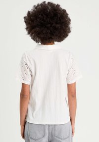 Chemise blanche à manches courtes avec un corps texturé, détails de broderie œillets sur les manches et un col. Coupe décontractée avec un ourlet droit.