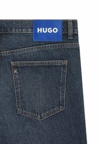 Ciemnogranatowe jeansy z denimu z niebieską skórzaną naszywką z logo "HUGO". Widoczna tylna kieszeń z delikatnym przeszyciem.