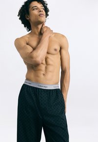 Čierne boxerské nohavičky s šedým opaskom, na ktorom je text "Calvin Klein". Zdá sa, že tkanina je hladká a má geometrický vzor.