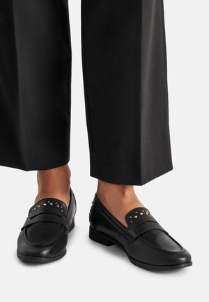 Czarne skórzane mokasyny z drobnymi złotymi ćwiekami, noszone na bosych stopach z czarnymi, skróconymi szerokimi spodniami.