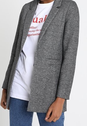 Person iført en grå tekstureret blazer over en hvid t-shirt med rød tekst og blå jeans, står mod en ensfarvet baggrund.