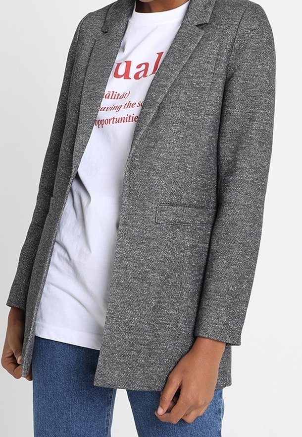 Personne portant un blazer gris texturé sur un t-shirt blanc avec un texte rouge et un jean bleu, debout devant un fond uni.